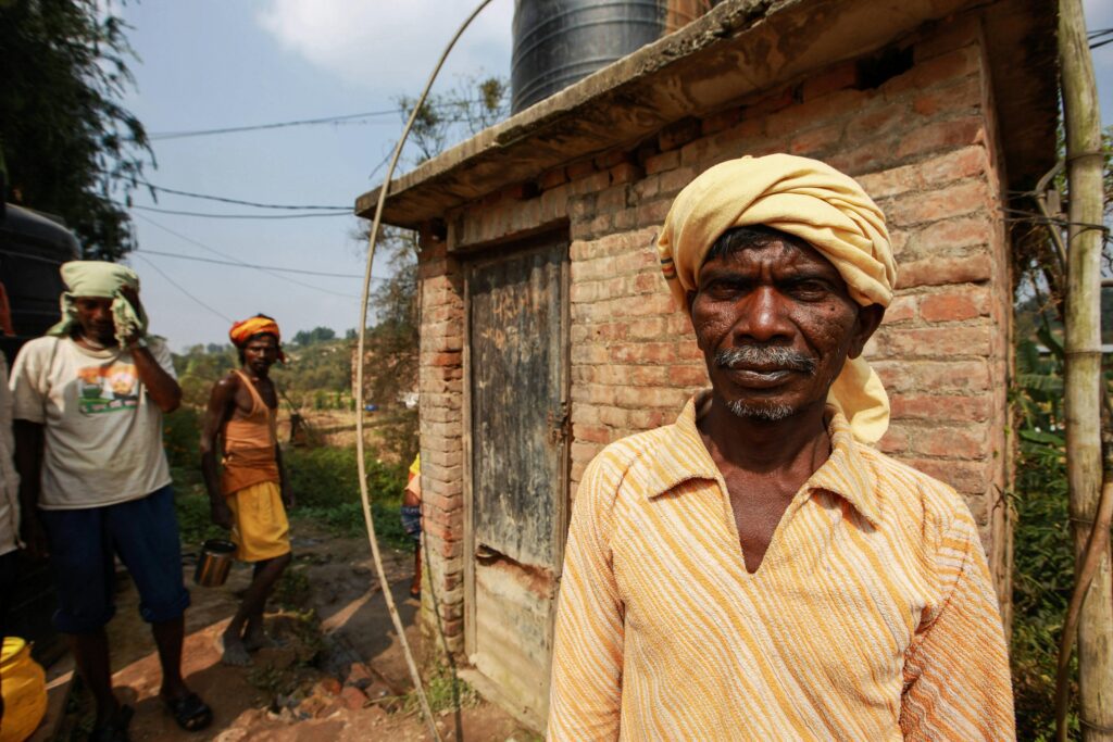 Elderly man in rural India wearing a turban, standing by a brick structure.